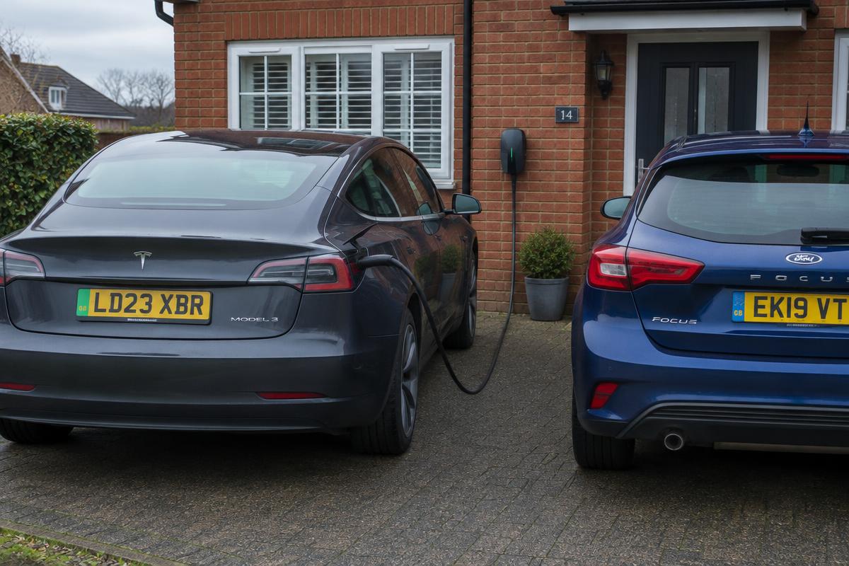 Electric car charging from a wall charger on a home driveway next to a petrol hatchback